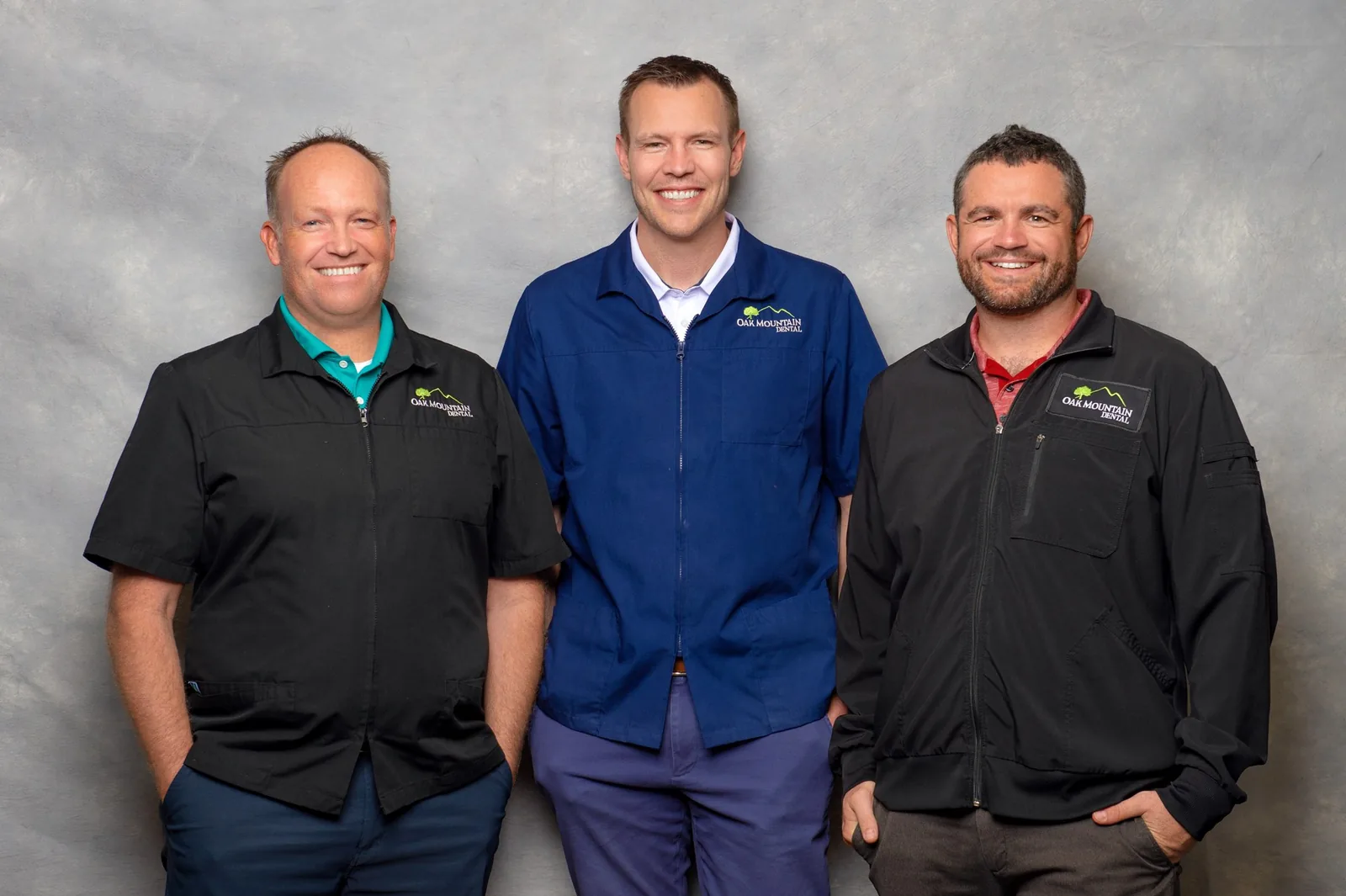 Three smiling dental professionals wearing Oak Mountain Dental uniforms in front of a gray backdrop.
