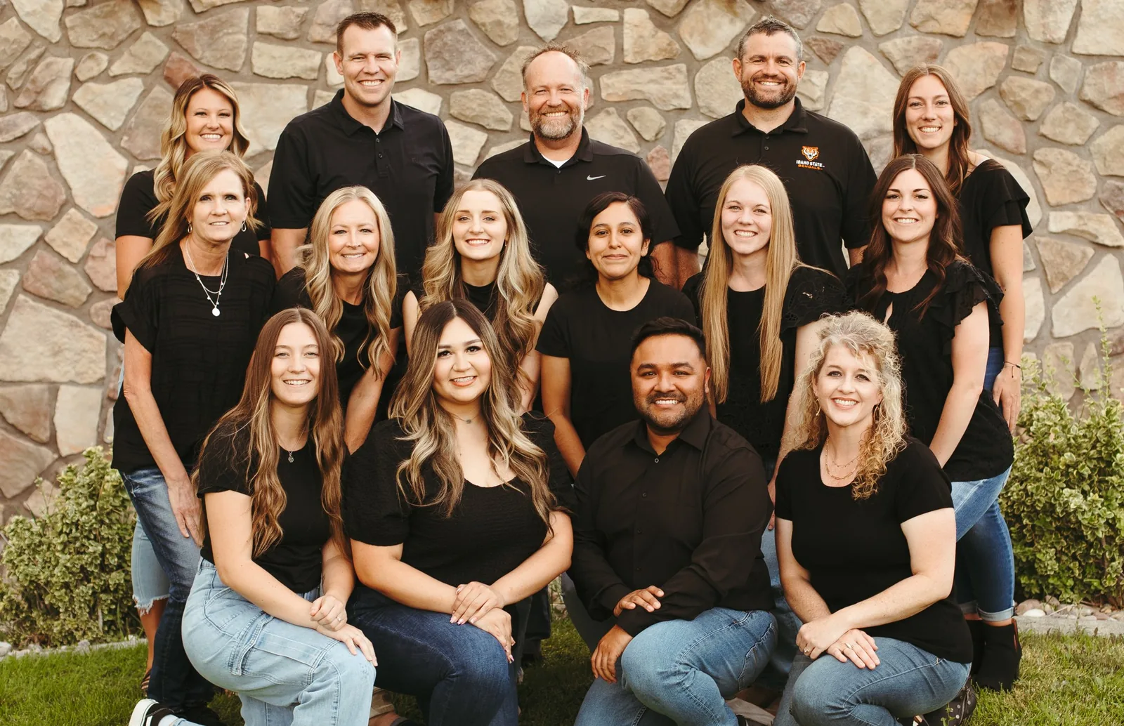 A group of dental team members smiling outdoors in front of a stone wall, wearing black tops and jeans.